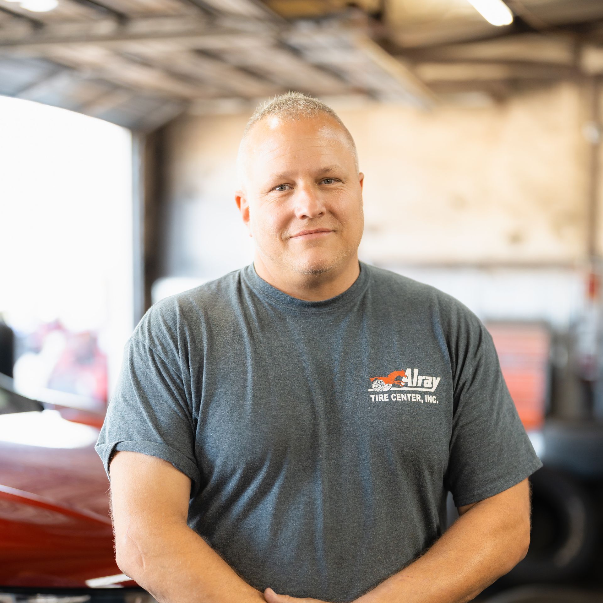 Man standing in a garage beside a red car, wearing a gray T-shirt and smiling.