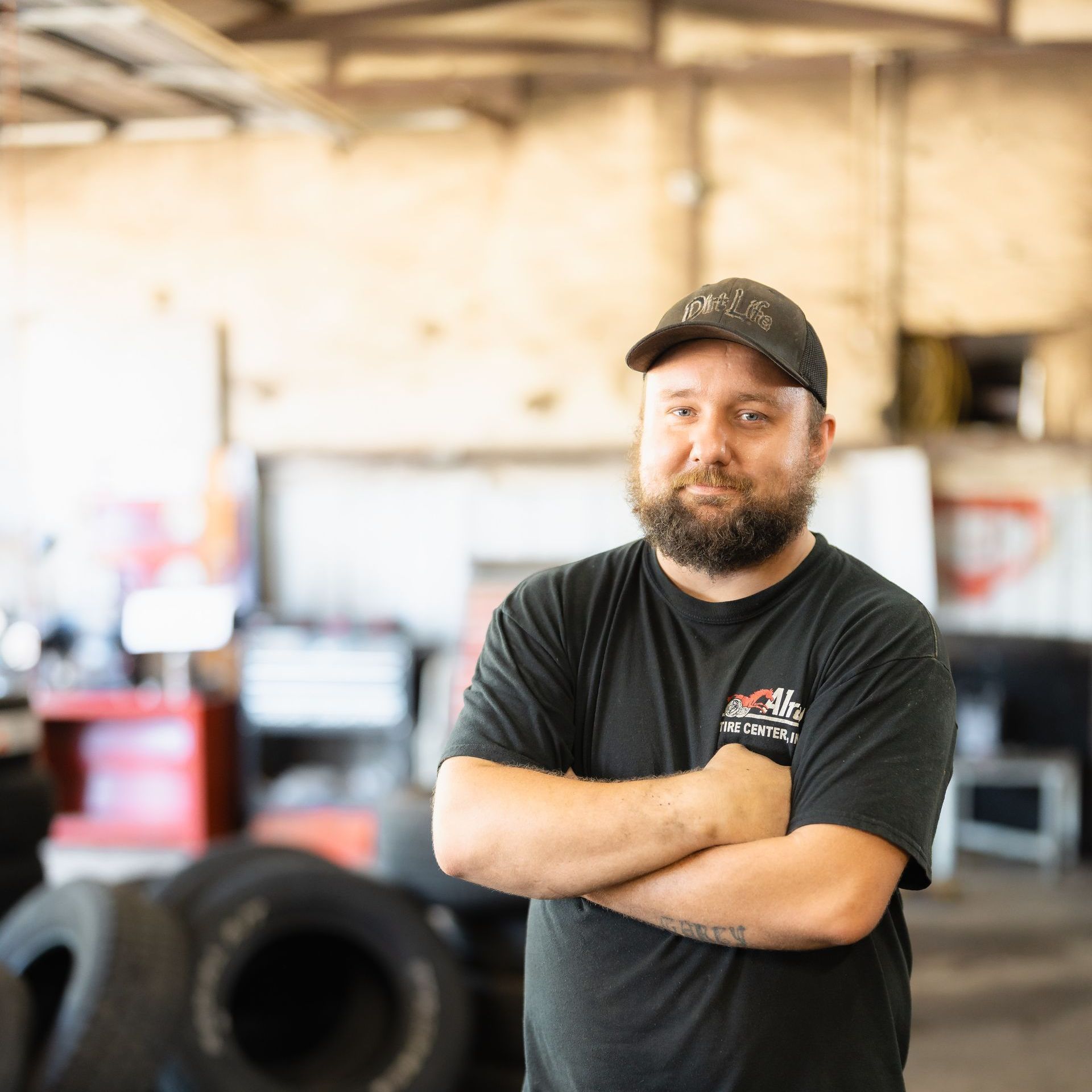 Man standing with arms crossed in a black cap and T-shirt inside a garage or workshop