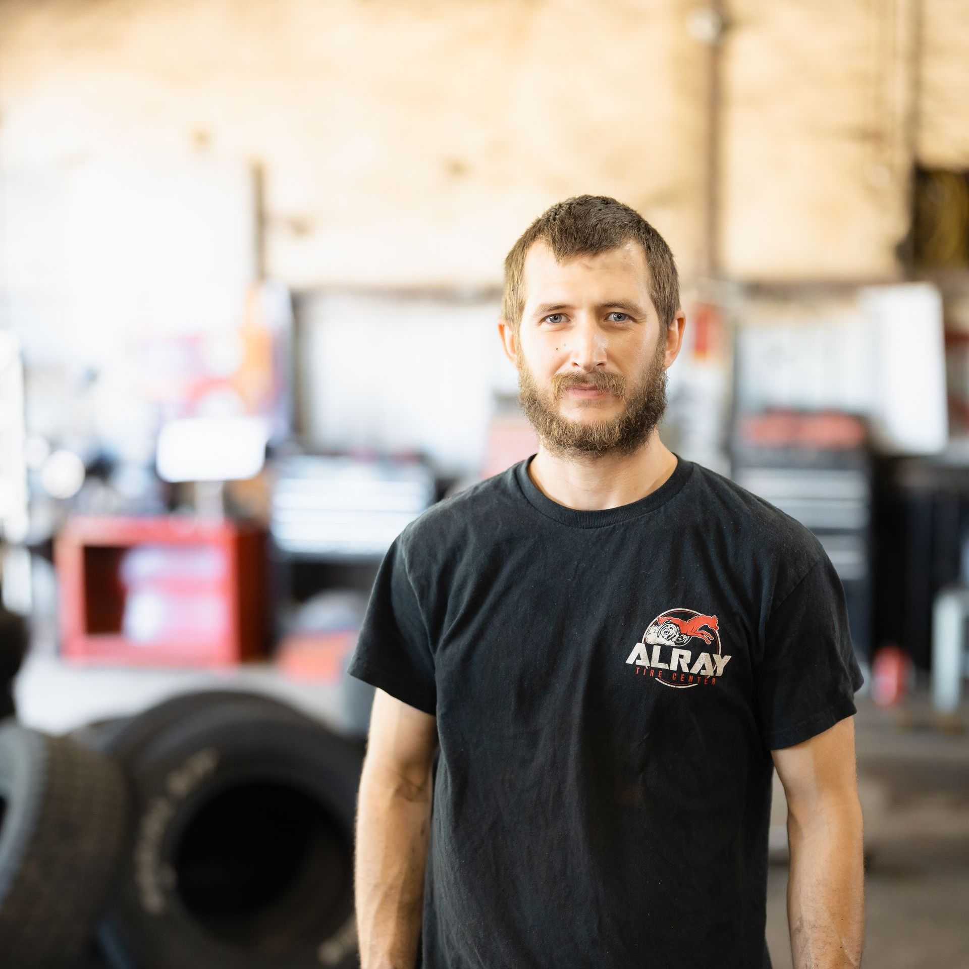 Man standing in a workshop, wearing a black T-shirt, with tires and equipment in the background