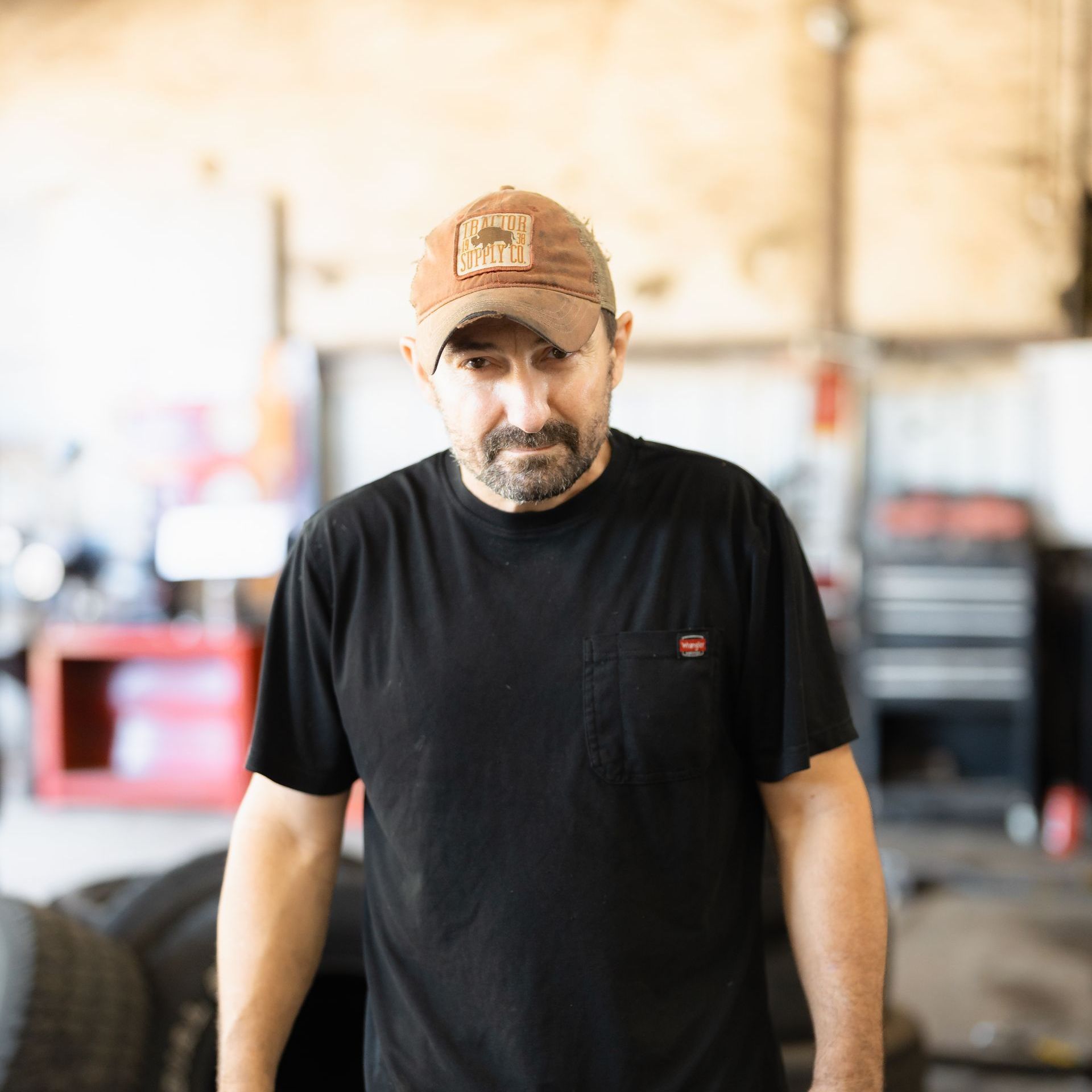 Man in a black T-shirt and tan cap standing in a workshop, looking down seriously