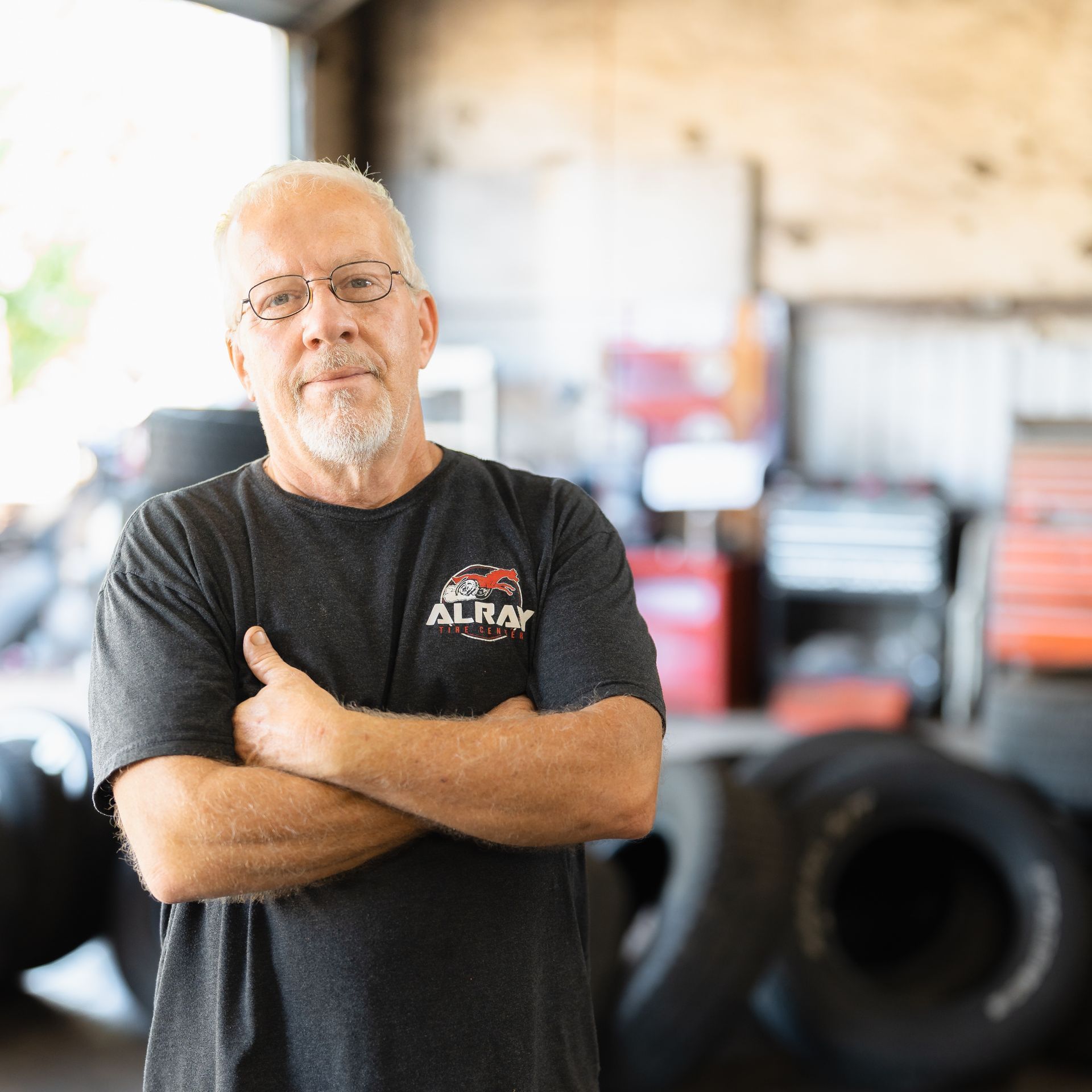 Man standing with arms crossed in a garage, with tires and equipment blurred in the background.