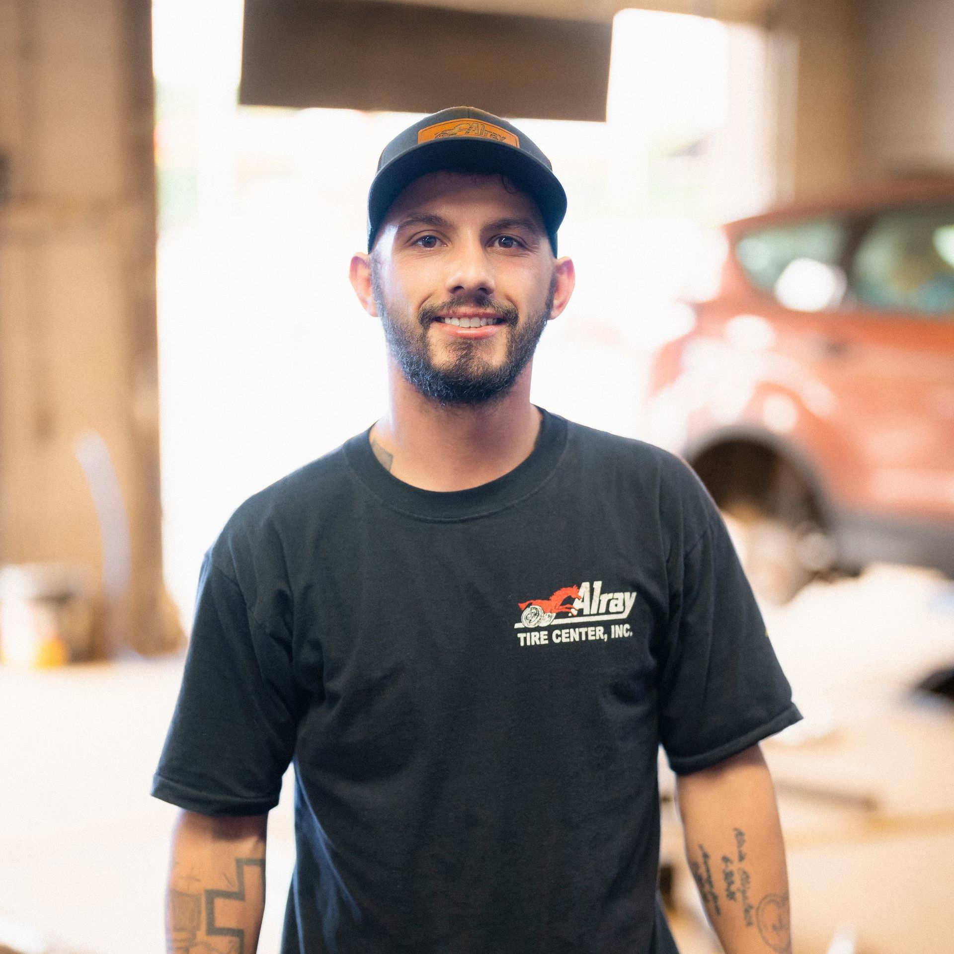 Man in black cap and T-shirt standing in a workshop, smiling at the camera.
