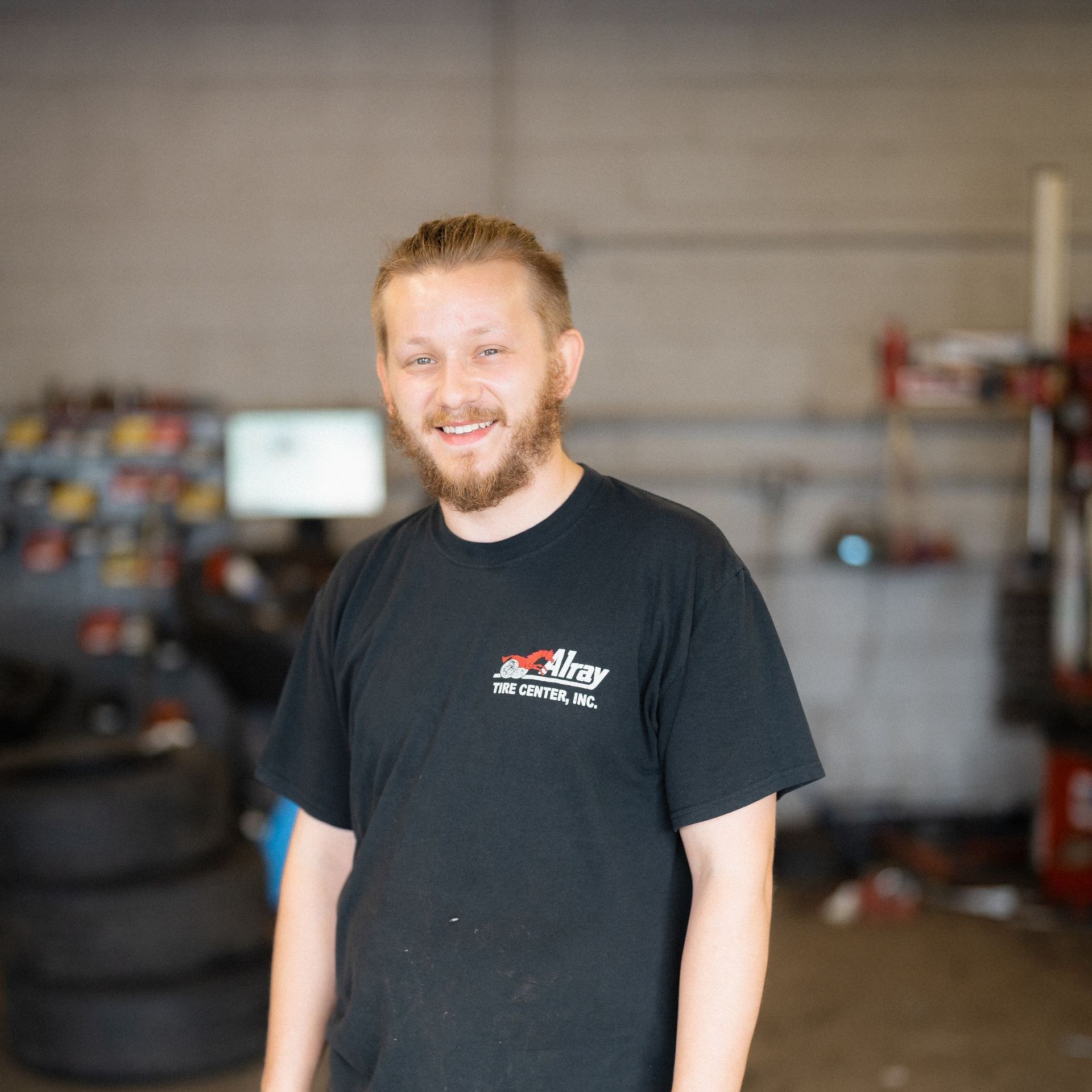 Smiling person in a black T-shirt standing in a garage or workshop with tires and equipment in the background