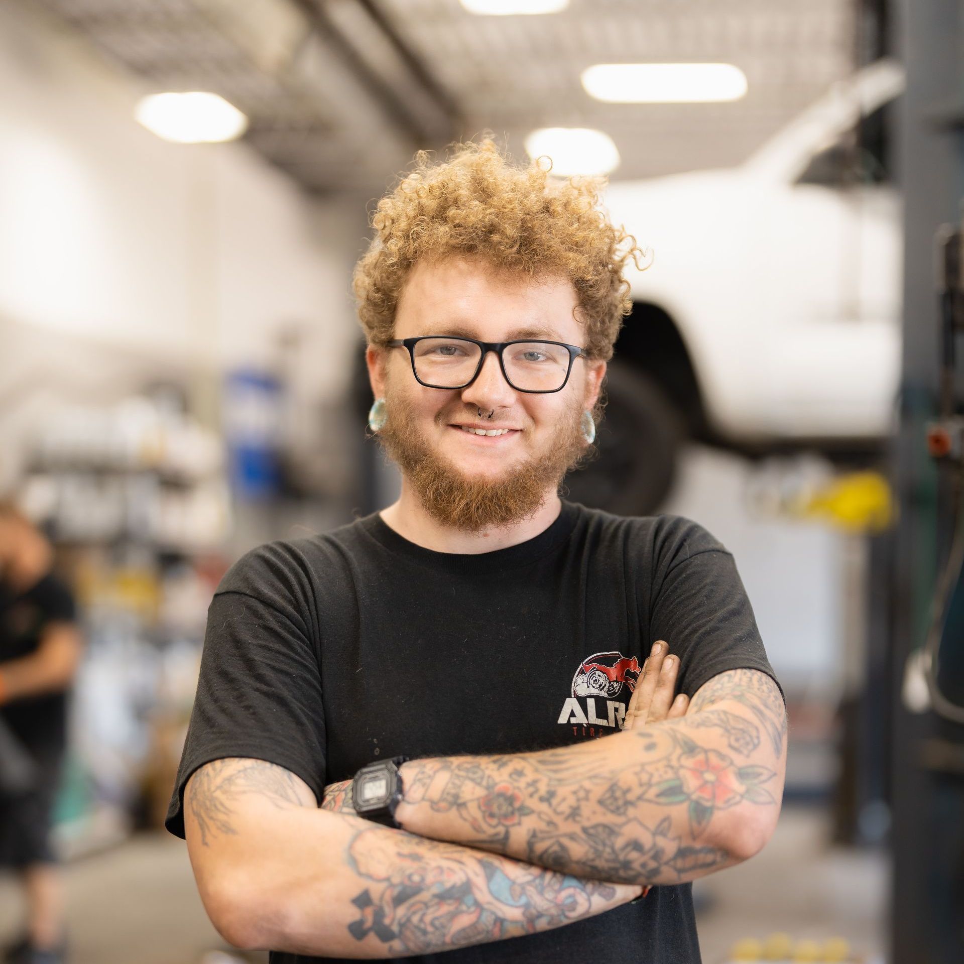 Smiling person with curly hair, glasses, and tattooed arms standing with crossed arms in a workshop