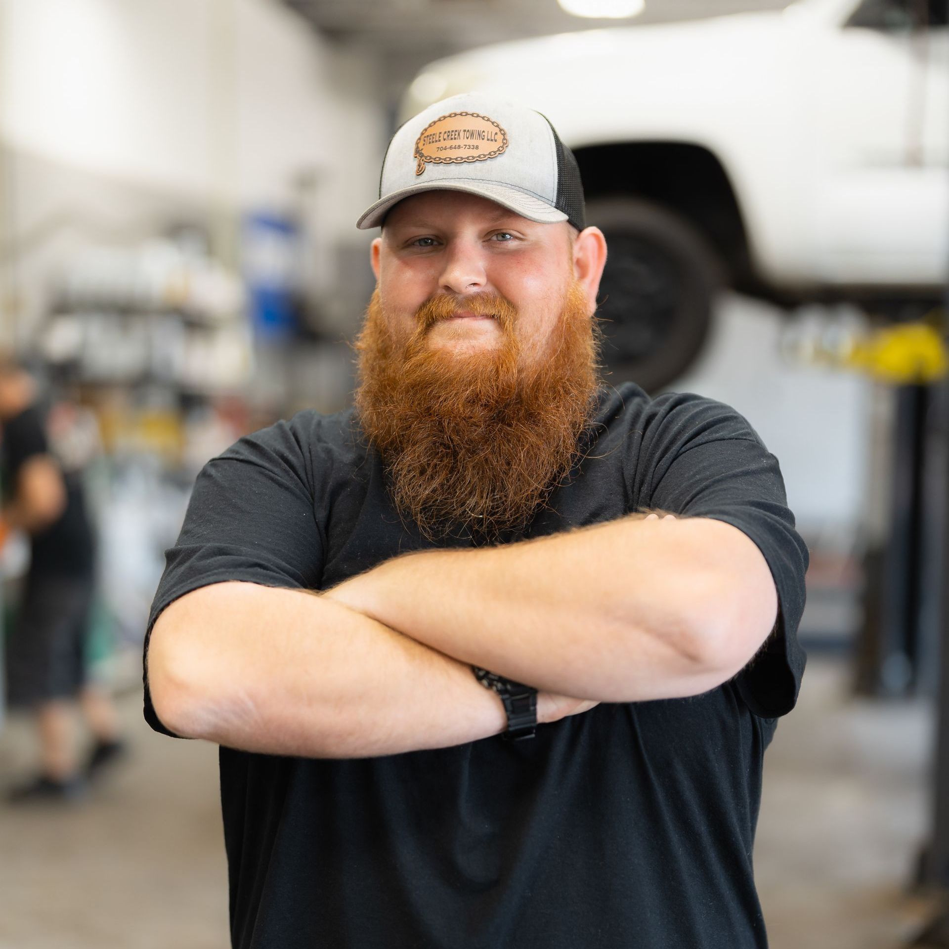 Smiling bearded person with crossed arms in a garage, wearing a cap and black shirt.