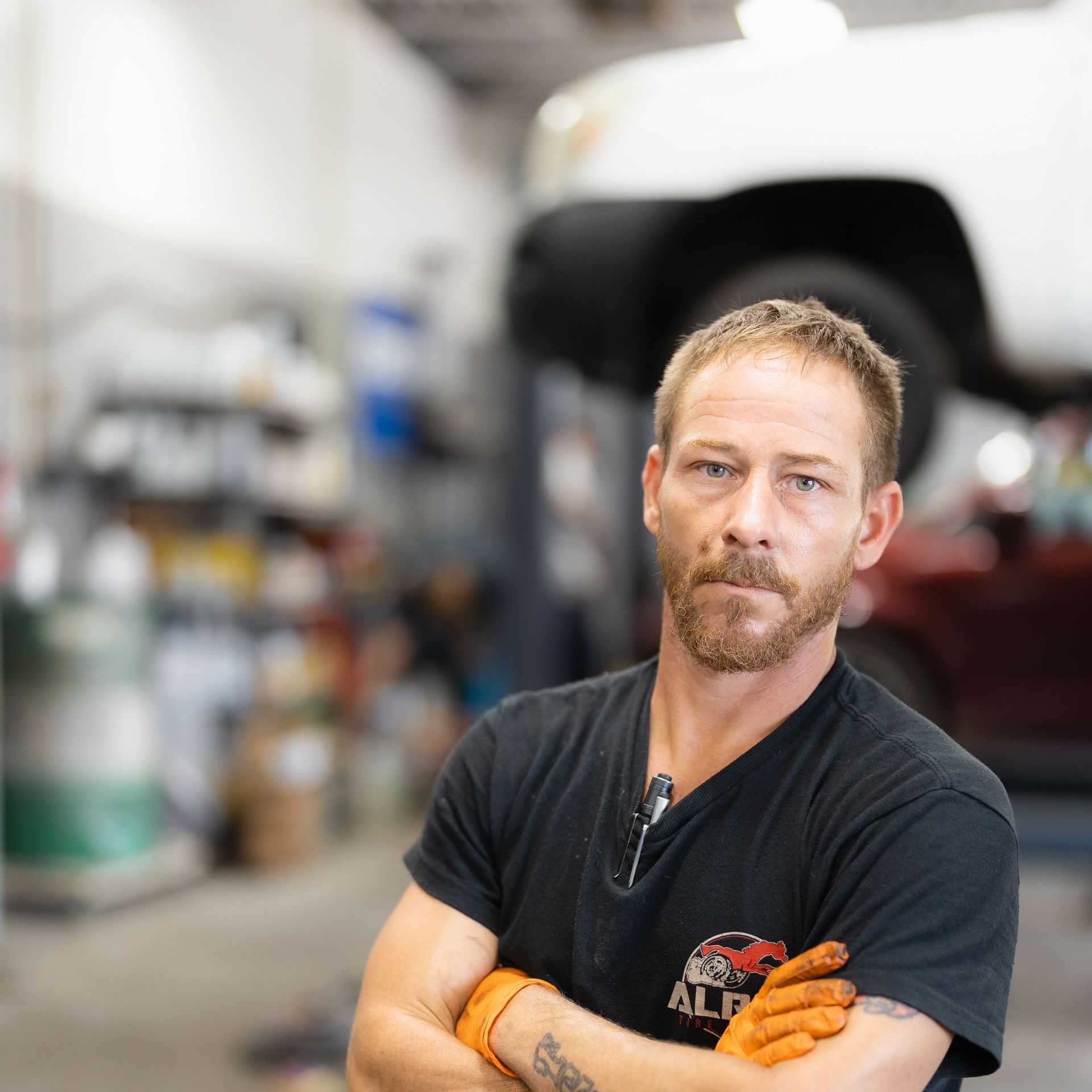 Mechanic with orange gloves standing in a garage, arms crossed, with a car in the background