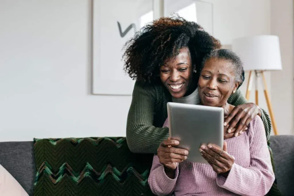 Two women smiling and looking at a tablet on a couch in a bright living room