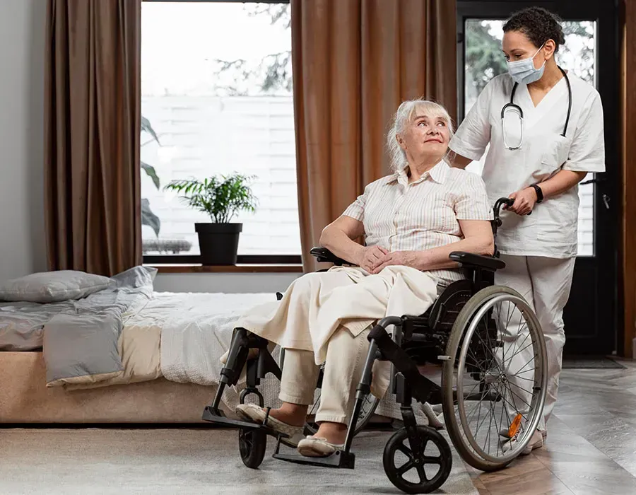 Nurse assisting a patient in a wheelchair beside a bed in a bright bedroom.