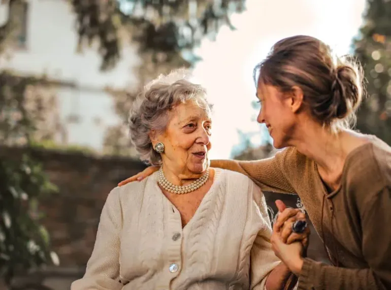 Two women smiling and chatting outdoors, one with an arm around the other