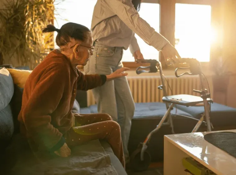 Person pushing a walker beside a seated woman in a sunlit living room