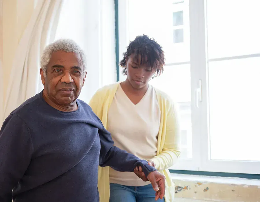 Two people standing by a bright window, one supporting the other with a hand on the arm.