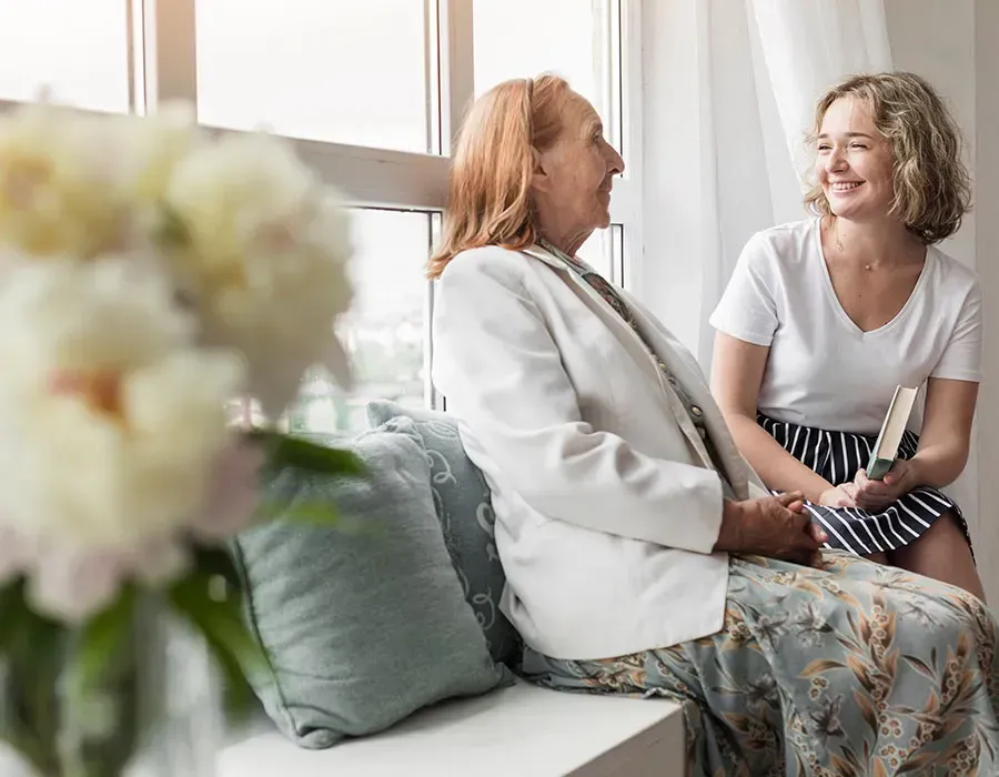 Two women smiling and talking on a sunlit couch beside a blurred bouquet and window.