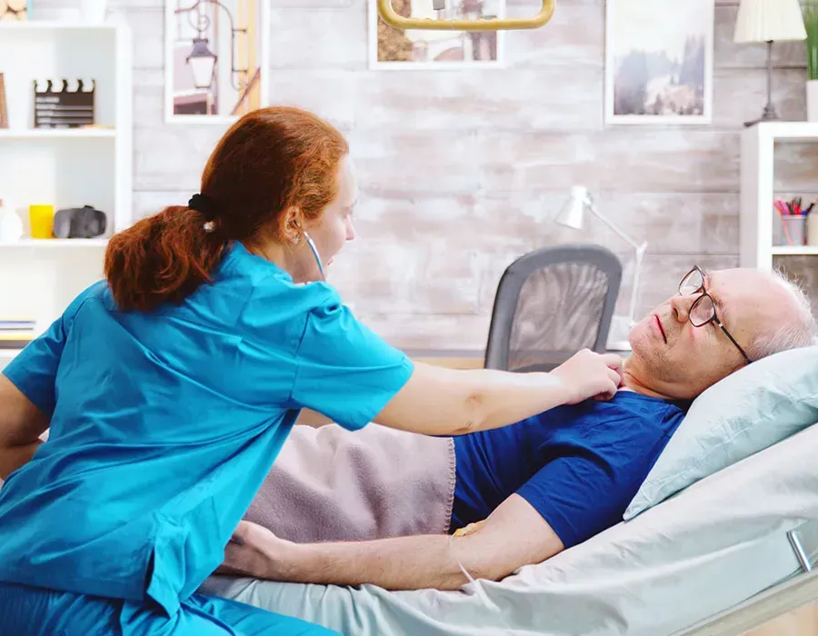 Caregiver adjusting an elderly patient’s shirt as he lies in bed in a bright home care room