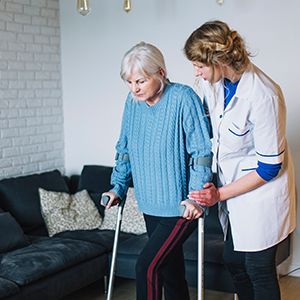 Caregiver helping a woman with a cane walk indoors beside a couch