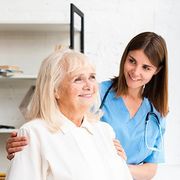 Smiling nurse in blue scrubs with stethoscope supporting an older patient in a bright clinic