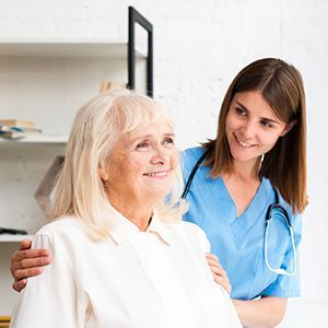 Smiling nurse in blue scrubs with stethoscope supporting an older patient in a bright clinic