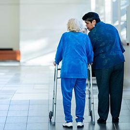 Two people walking in a bright hallway, one using a walker and the other assisting them