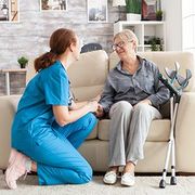 Caregiver in blue scrubs kneels beside smiling older woman on sofa with walker in living room