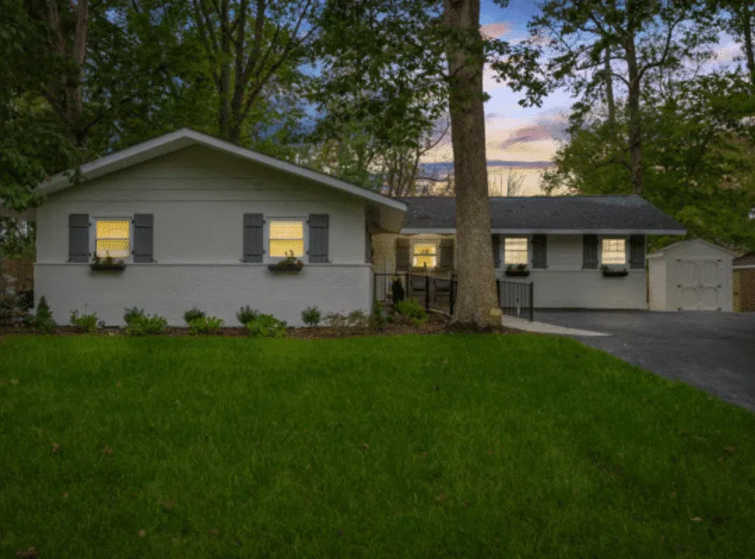 Suburban house at dusk with lit windows, large front lawn, and driveway beside tall trees.