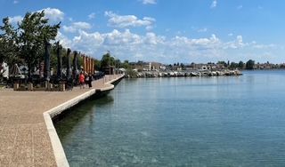 Curved waterfront promenade beside calm blue water under a sunny sky, with people and trees on the left