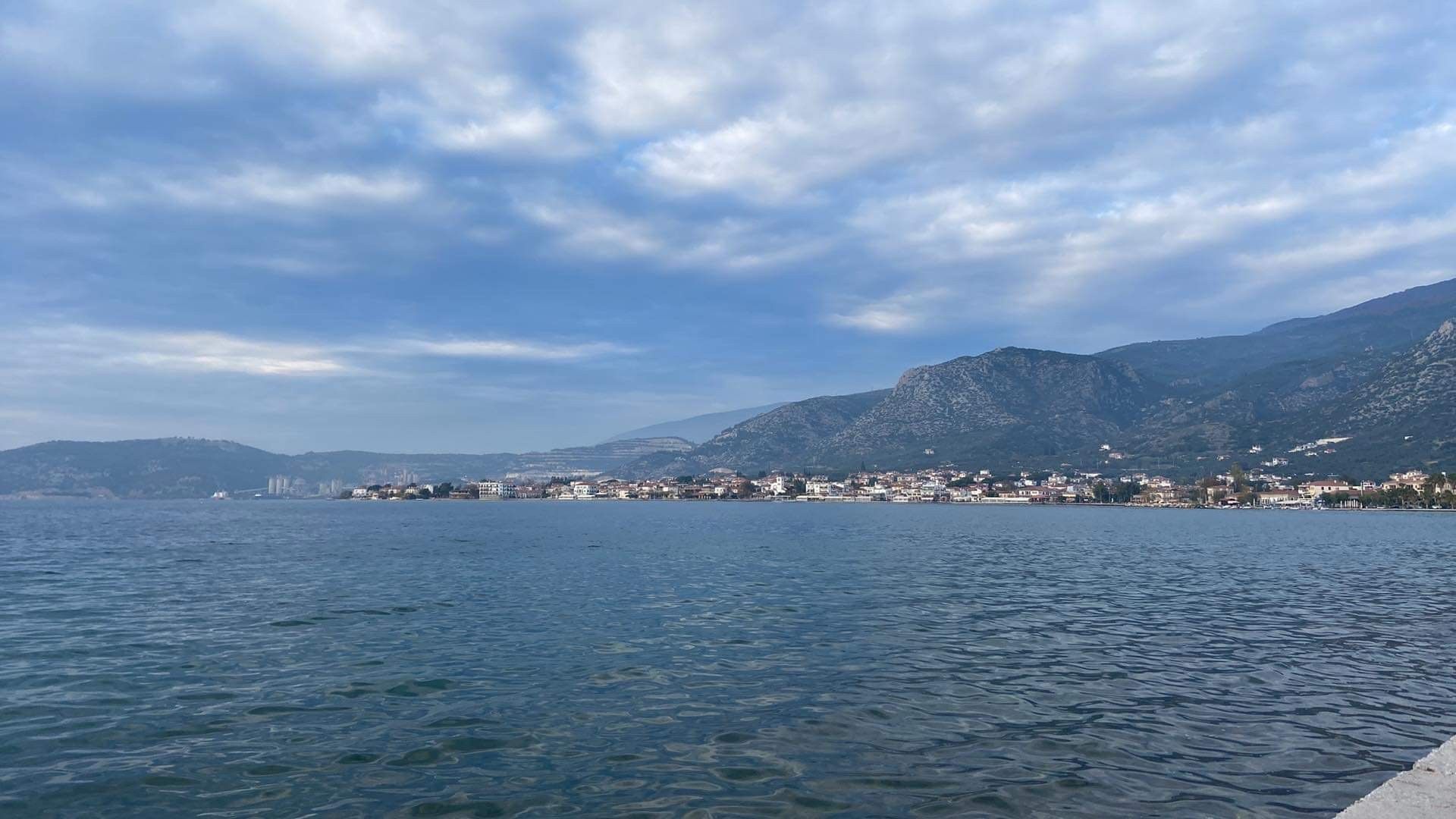 Calm blue sea beside a coastal town and mountains under a cloudy sky