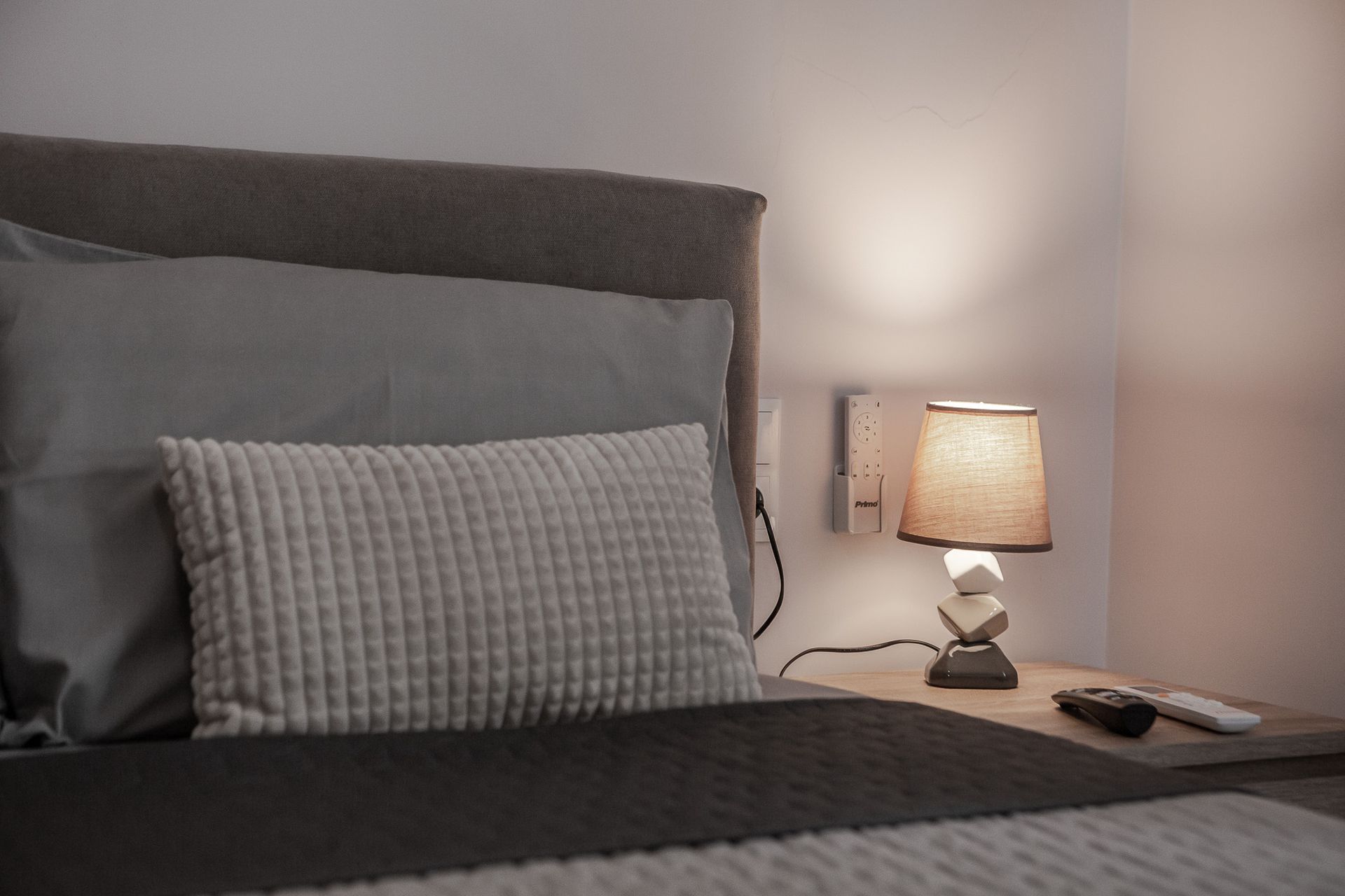 Neatly made bed with gray bedding beside a nightstand and lit table lamp in a softly lit bedroom.