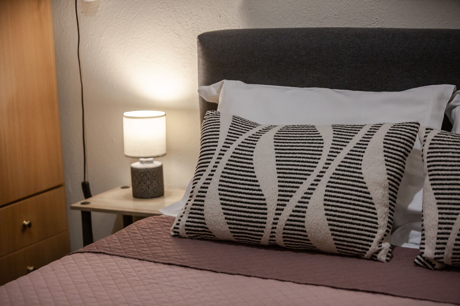 Cozy bedroom corner with lamp on nightstand, wood cabinet, and bed with patterned pillows and mauve blanket