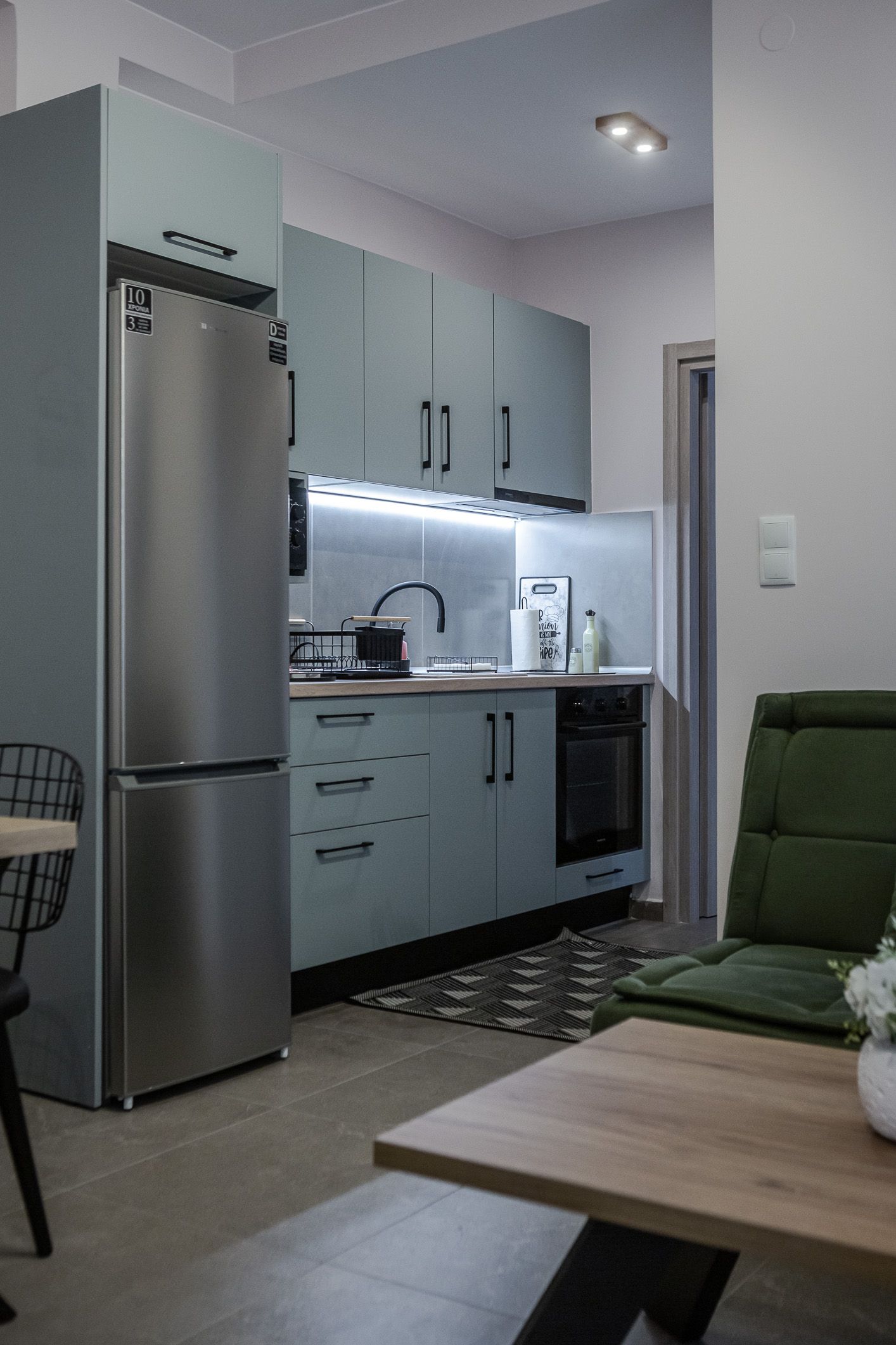 Compact kitchen with stainless steel fridge, white cabinets, and a green chair beside a coffee table