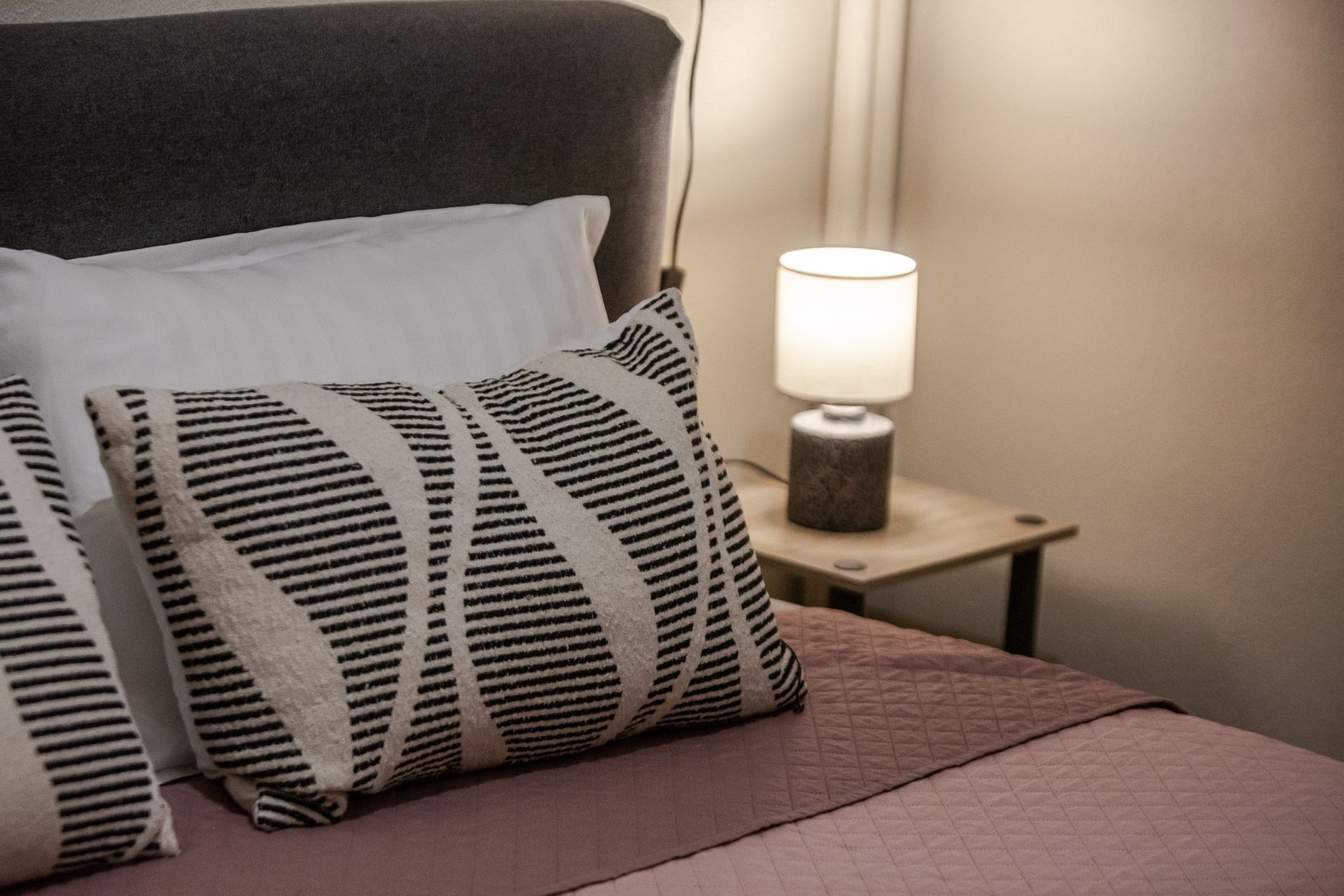 Bedroom with neatly made bed, patterned pillows, and a lit bedside lamp on a small nightstand