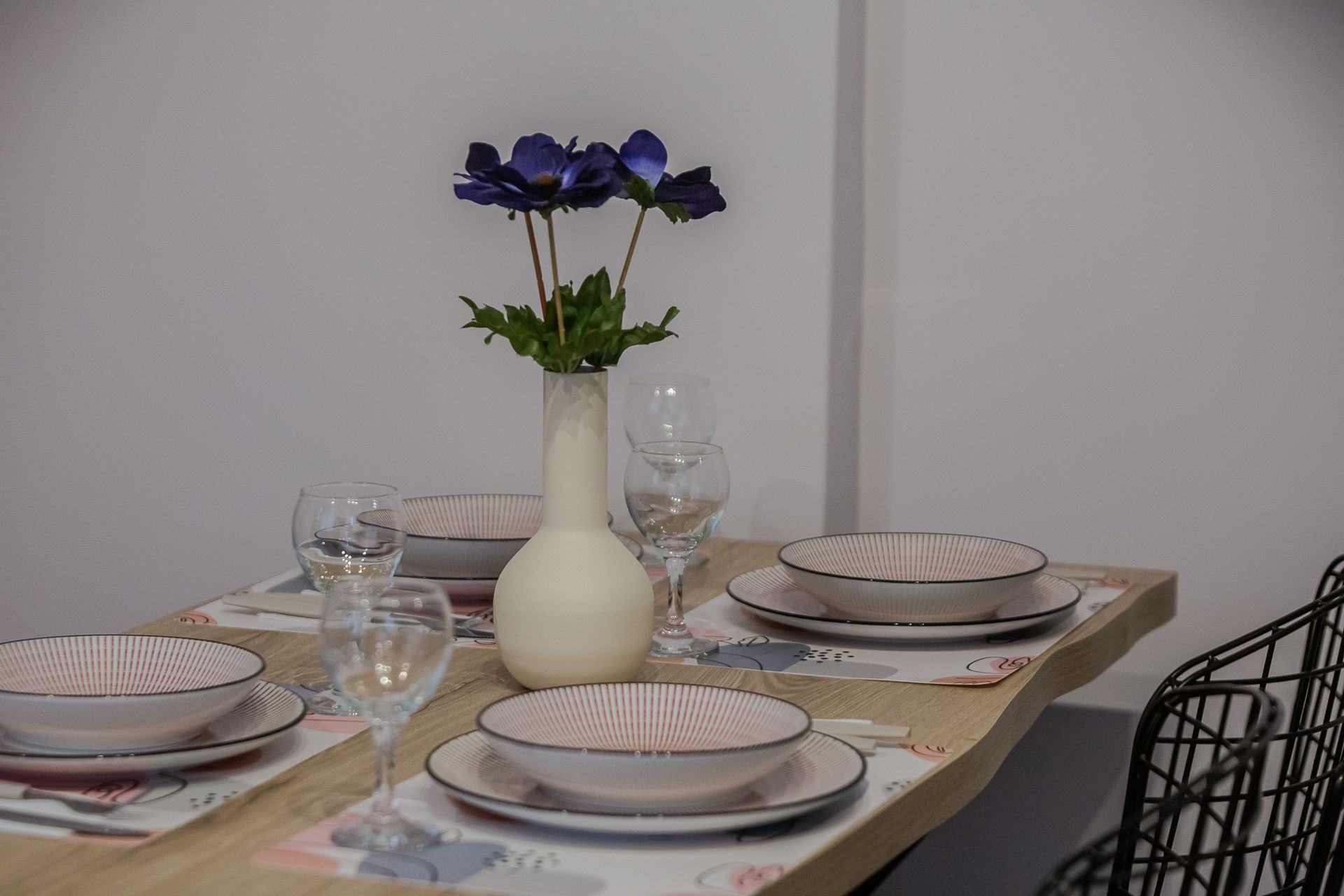 Dining table set for four with white dishes, glassware, and a vase of purple flowers.