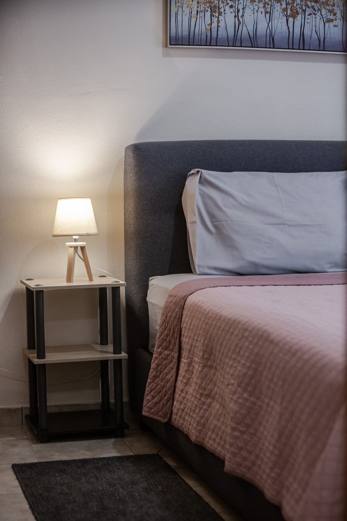 Small bedroom corner with lit lamp on a side table beside a gray bed and pink quilt.