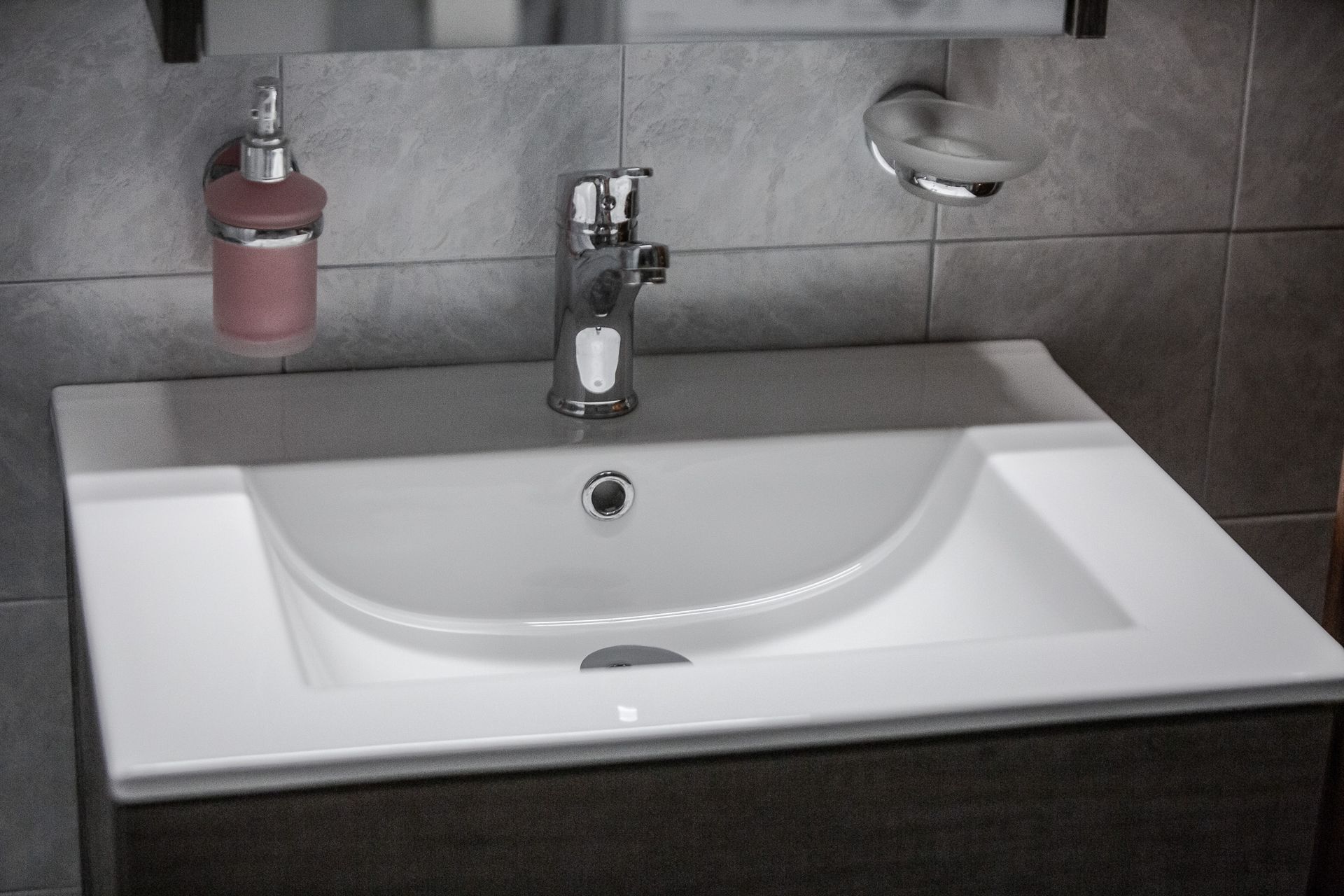 Modern white bathroom sink with chrome faucet and pink soap dispenser against gray tile wall