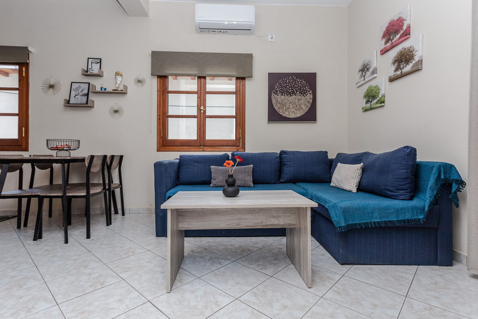 Bright living room with blue sofa, coffee table, wall art, and a small dining area by the window.