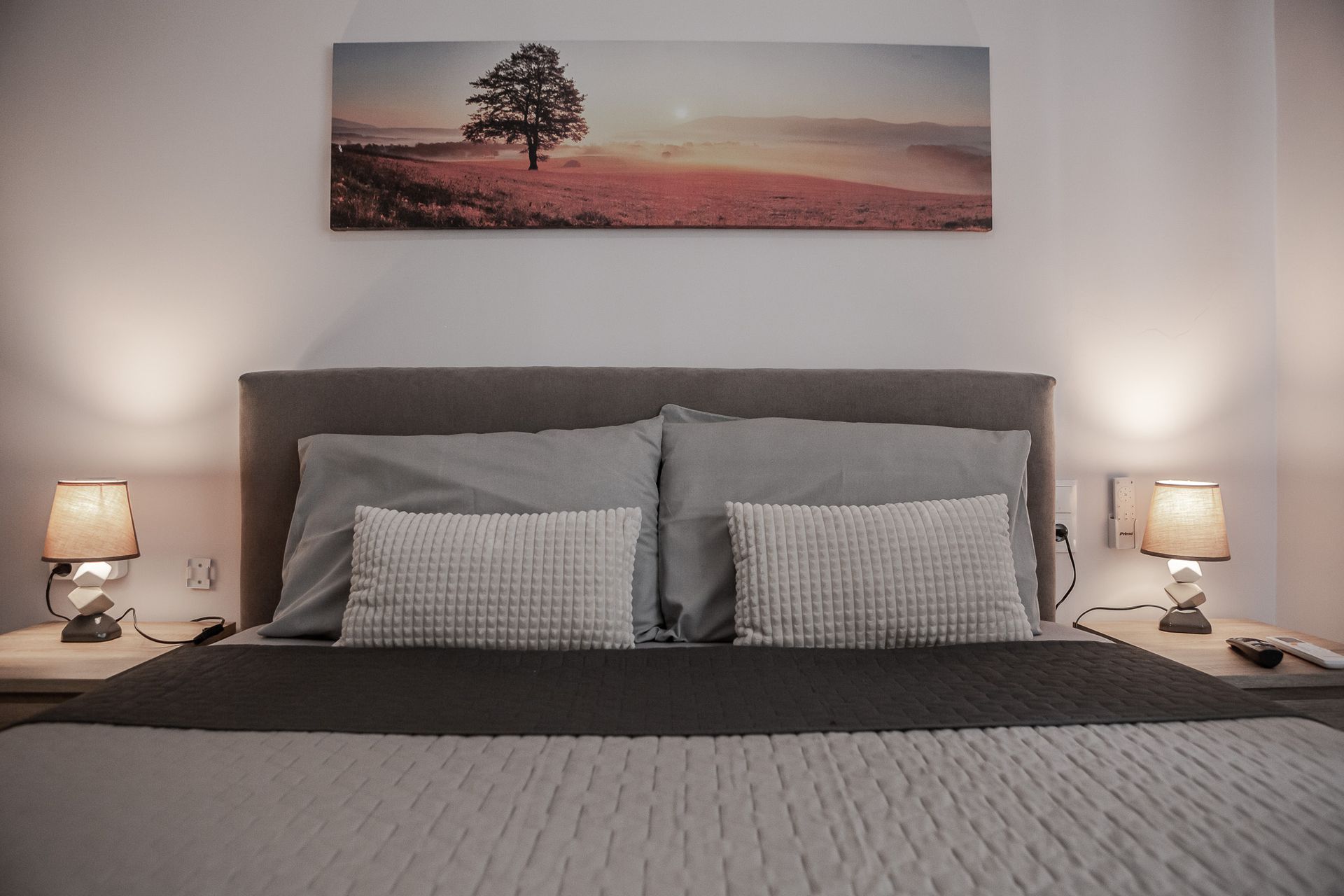 Neatly made bed with gray bedding, bedside lamps, and a landscape print above the headboard.