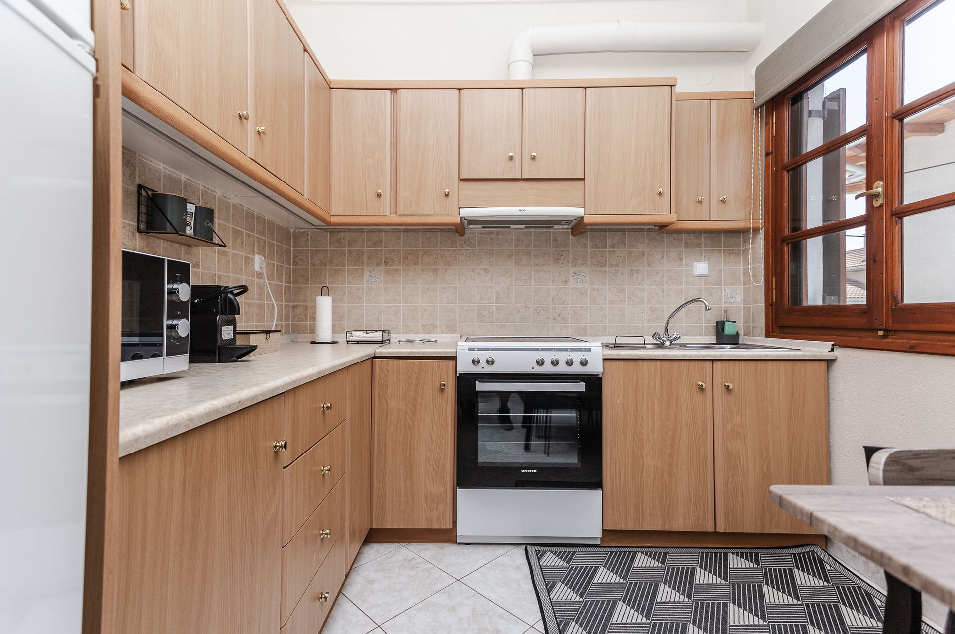 Small beige kitchen with wooden cabinets, white stove, countertop appliances, and a window above the sink