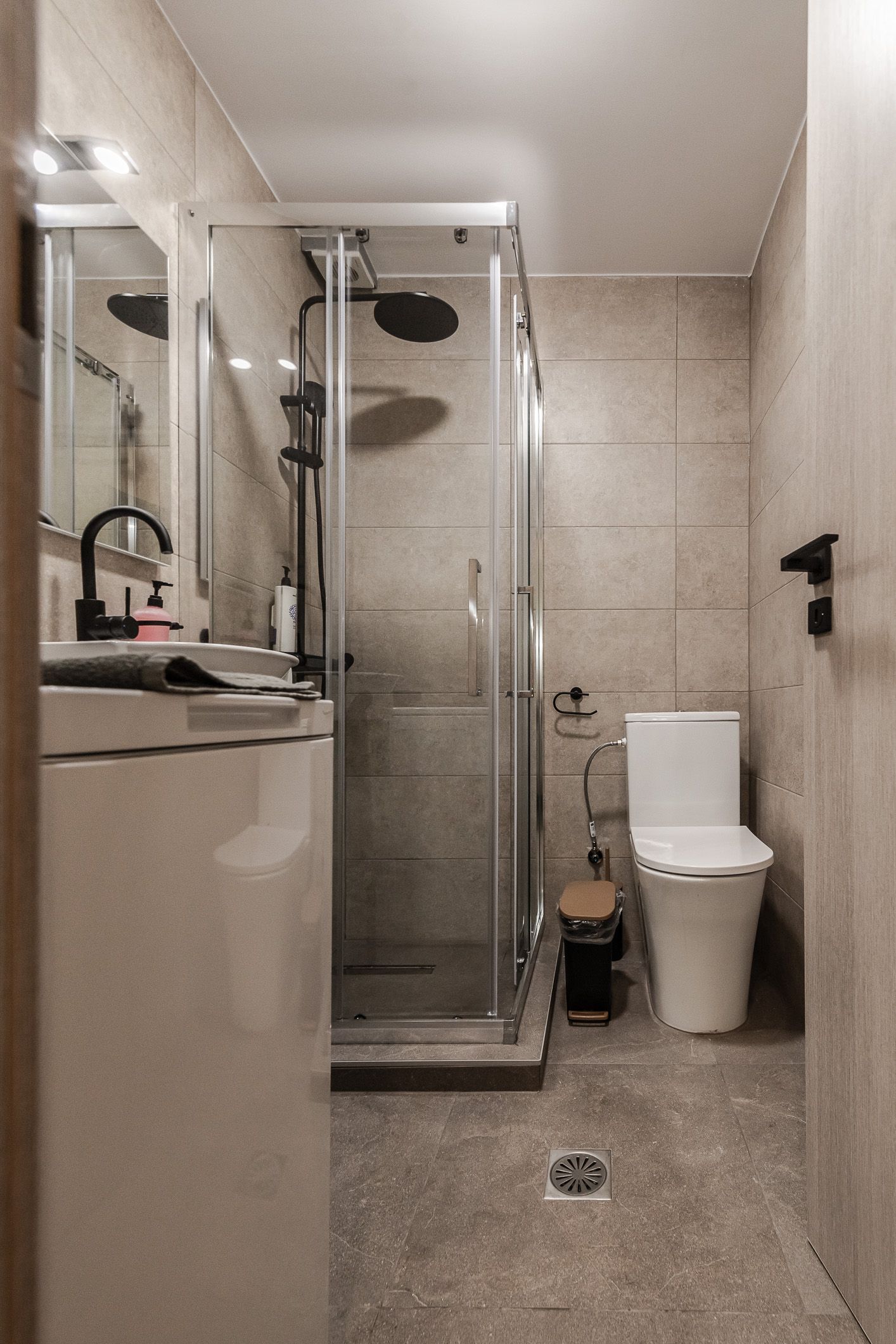 Narrow beige bathroom with glass shower stall, toilet, sink, and wall-mounted toilet paper holder