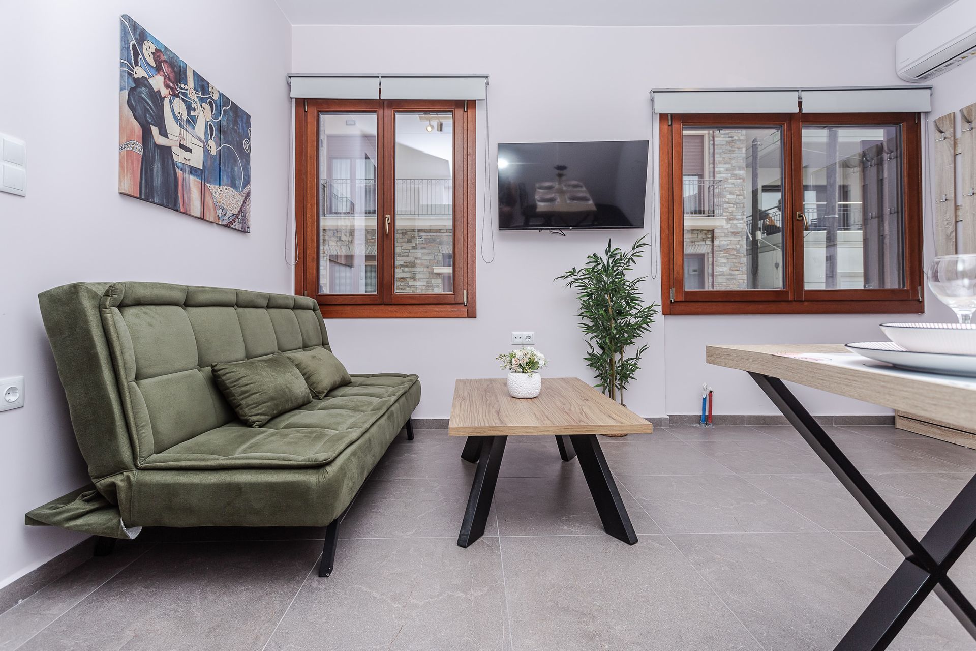 Cozy living room with green sofa, wooden coffee table, wall TV, and two windows on white walls.