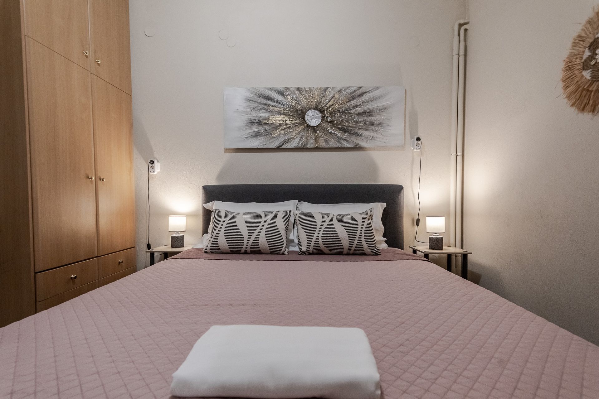 Cozy bedroom with a pink bedspread, patterned pillows, bedside lamps, and a wall art above the headboard