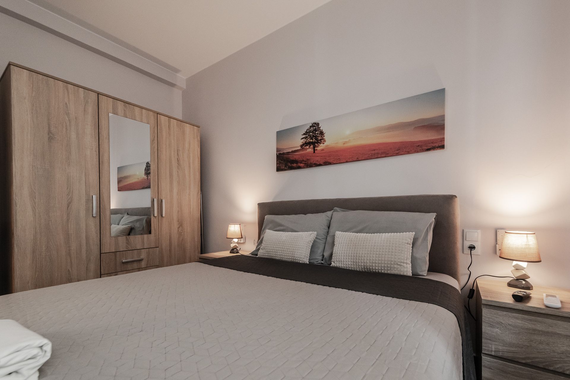 Modern bedroom with gray bed, wooden wardrobe, nightstands, and sunset wall art