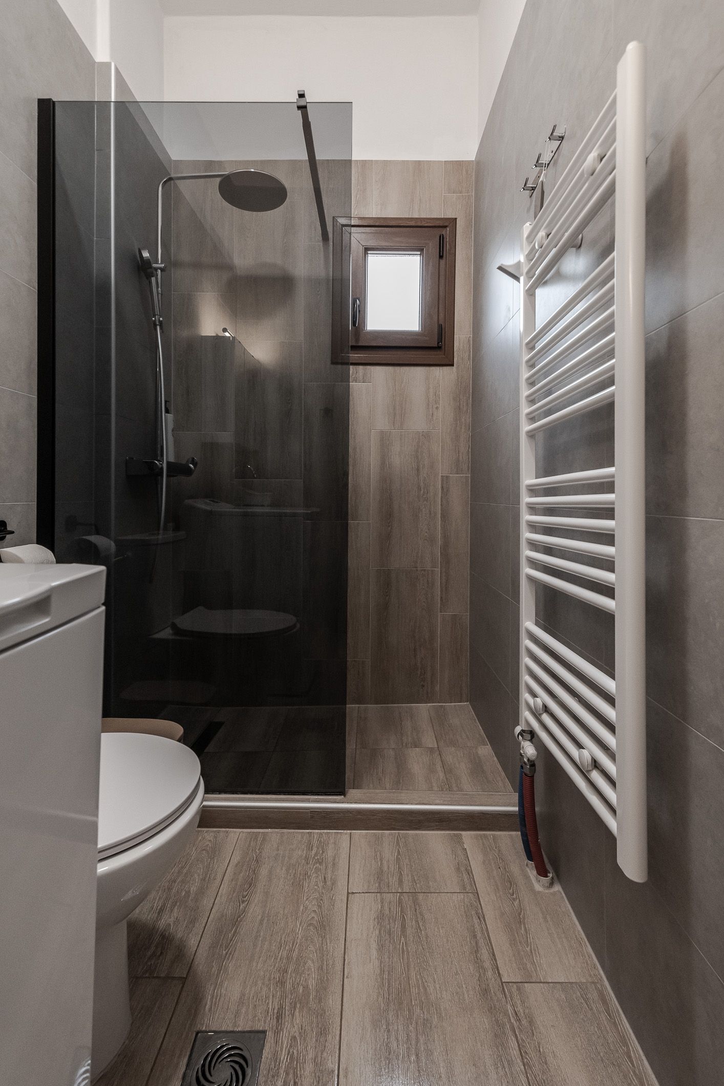 Small bathroom with a glass shower, toilet, towel radiator, and wood-look tile floor.