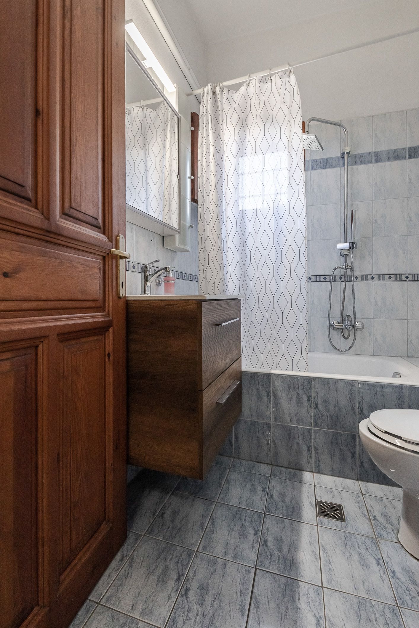 Bathroom with marble-look tile, glass shower, wooden vanity, and a partial toilet visible.