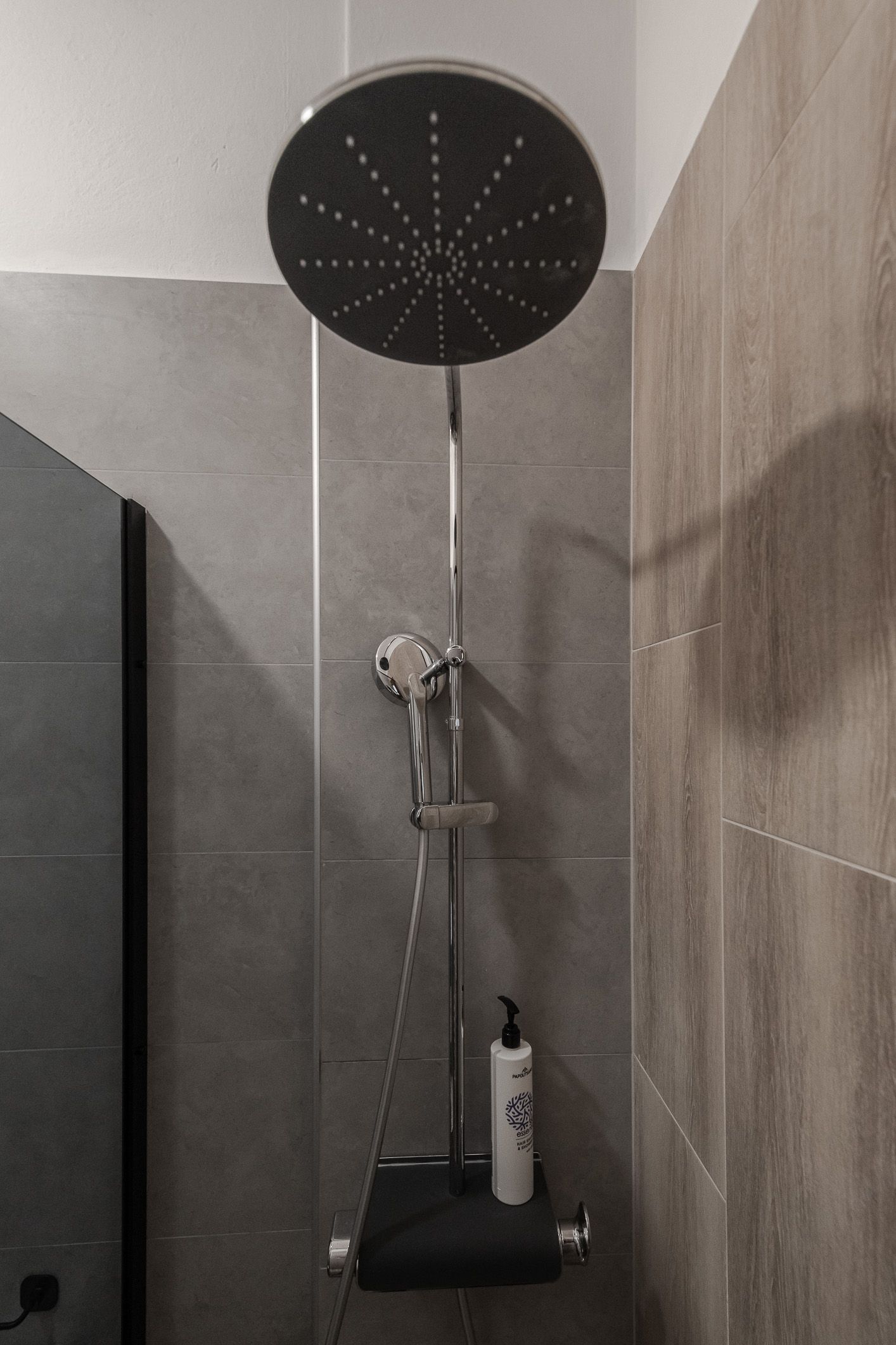 Bathroom shower with large round rain showerhead and black shelf holding toiletries.