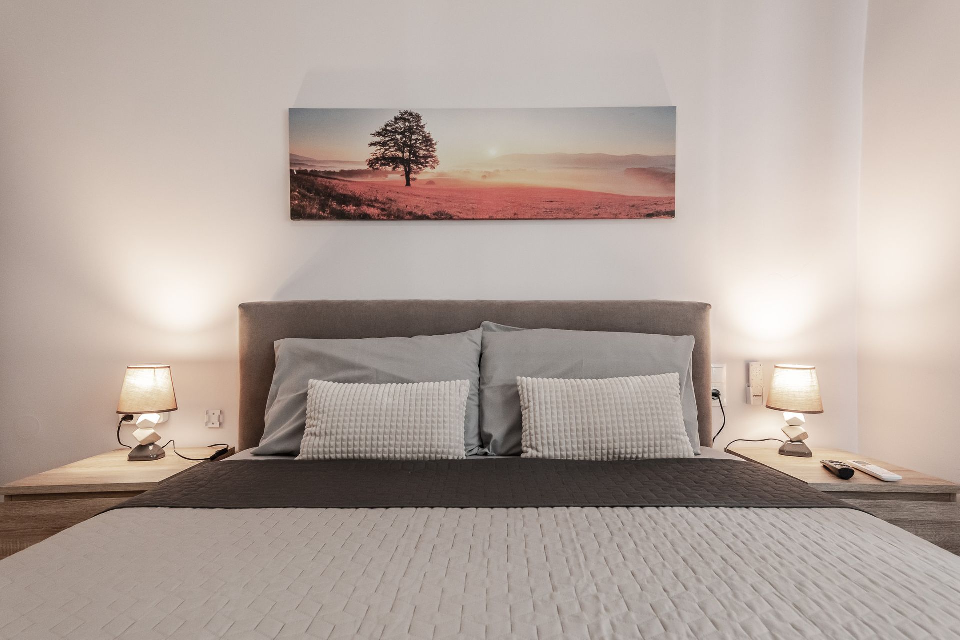 Cozy bedroom with gray bed, bedside lamps, and a landscape painting above the headboard.