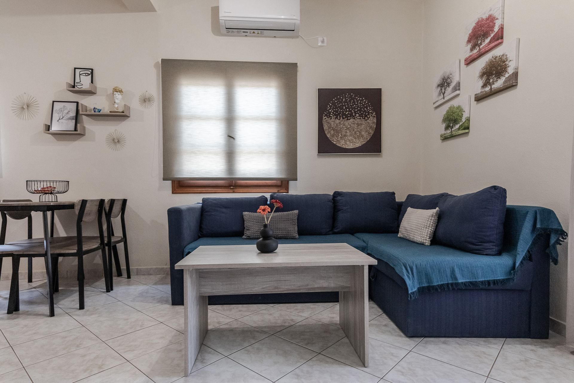 Cozy living room with a blue sectional, coffee table, wall art, and dining area near a window.