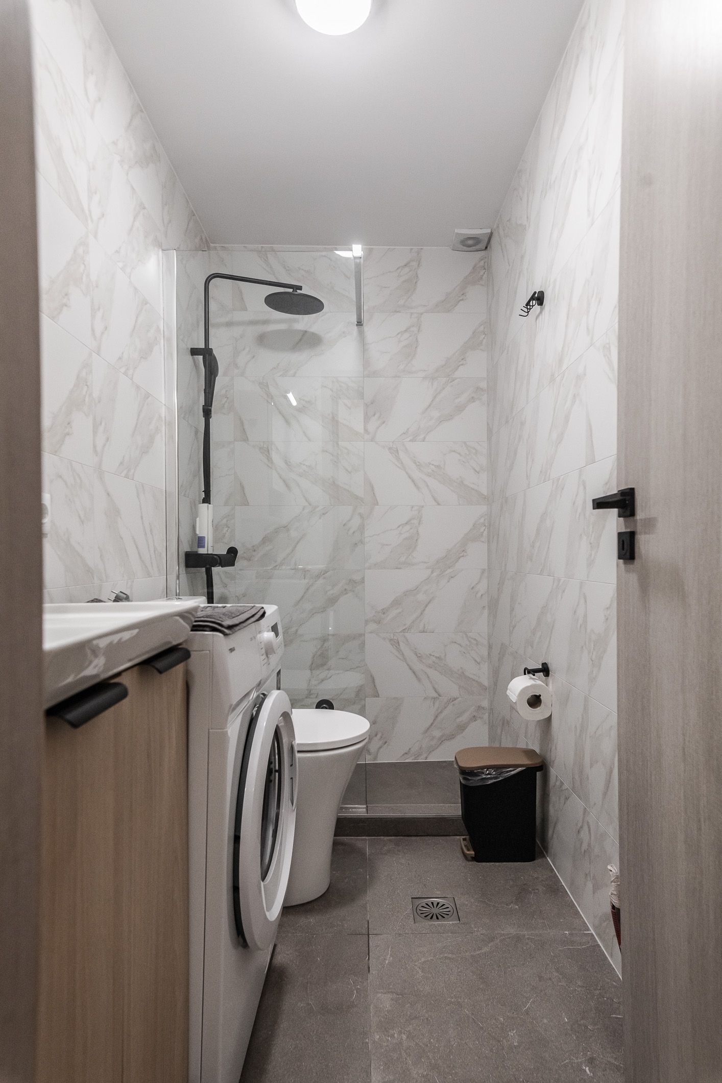 Narrow bathroom with toilet, shower, and white marble-patterned walls and floor.