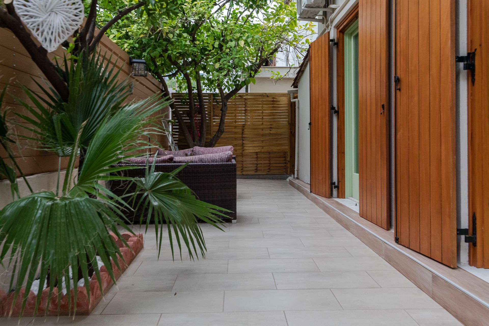Narrow courtyard with wooden doors, potted palms, and a wicker sofa along a tiled walkway.