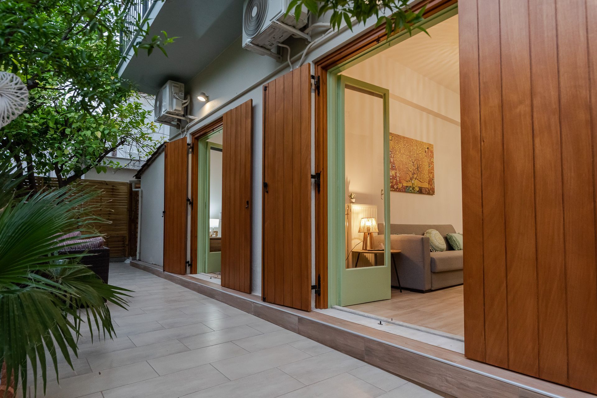 Modern home exterior with open wooden doors revealing a lit living room and patio walkway under trees