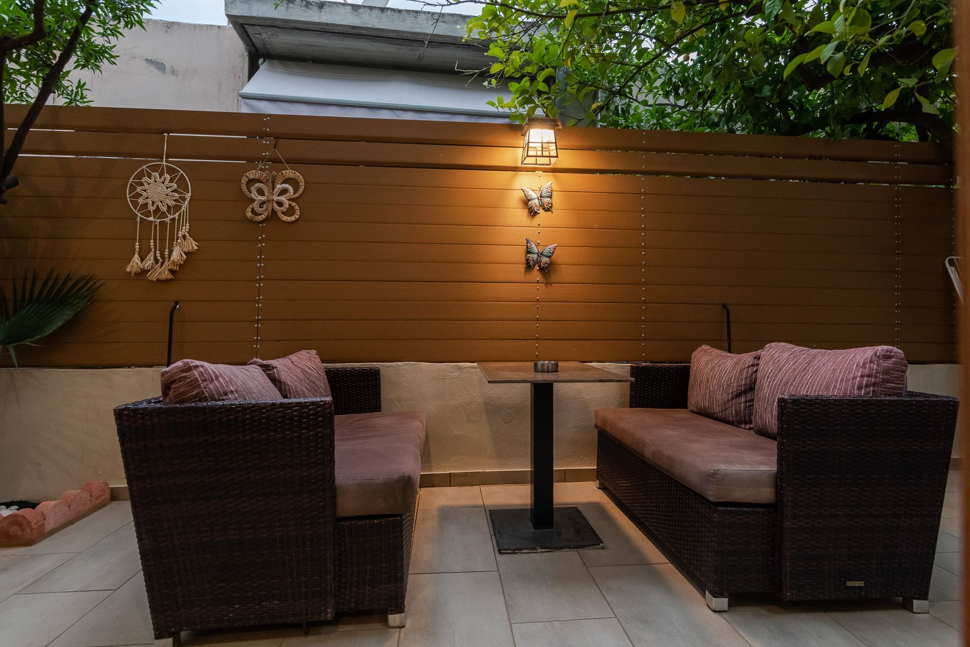 Outdoor patio with two wicker sofas, a small table, and warm wall lights against a wooden fence.