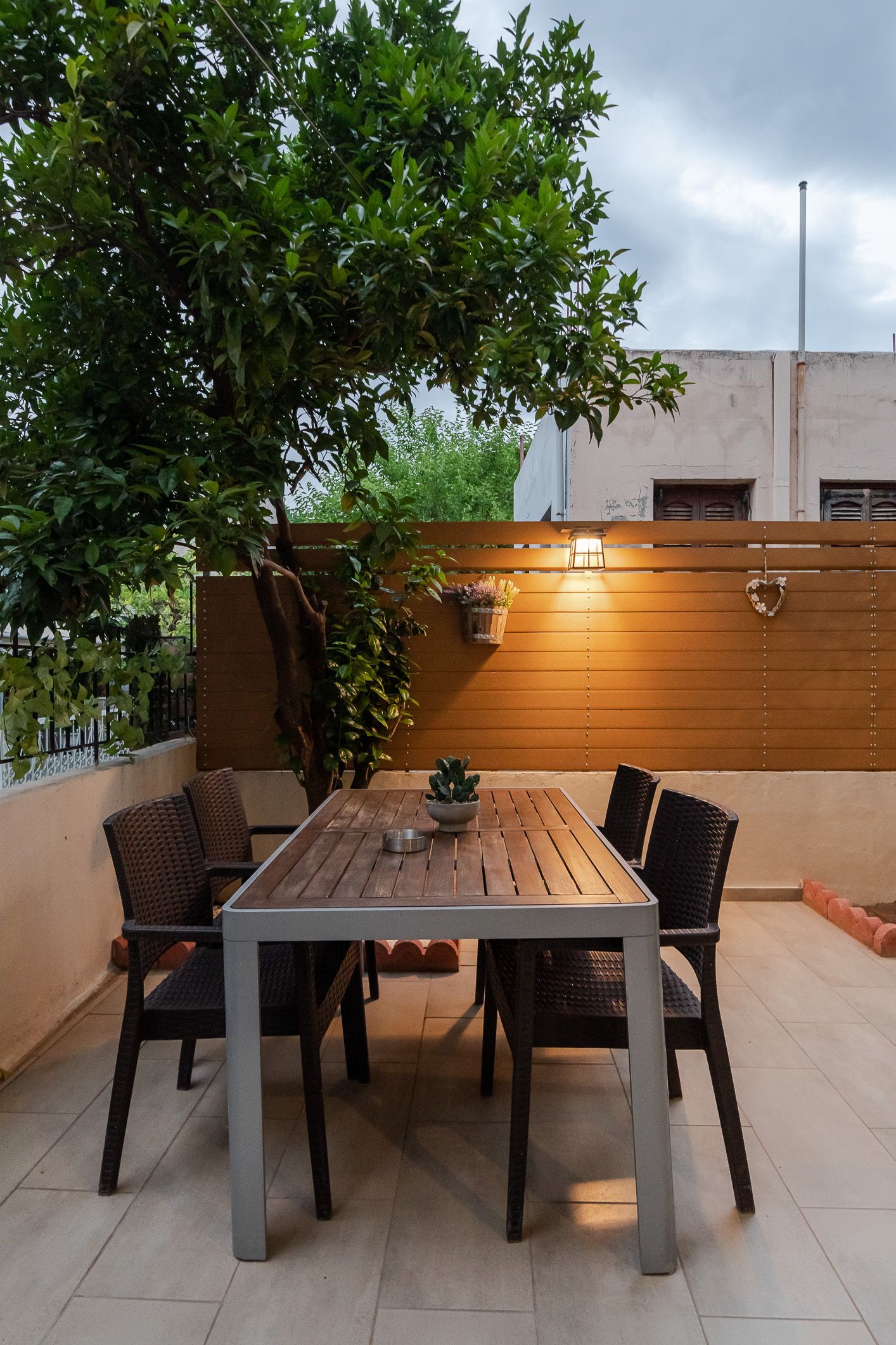 Outdoor patio with a wooden table, chairs, and a tree beside a lit fence at dusk