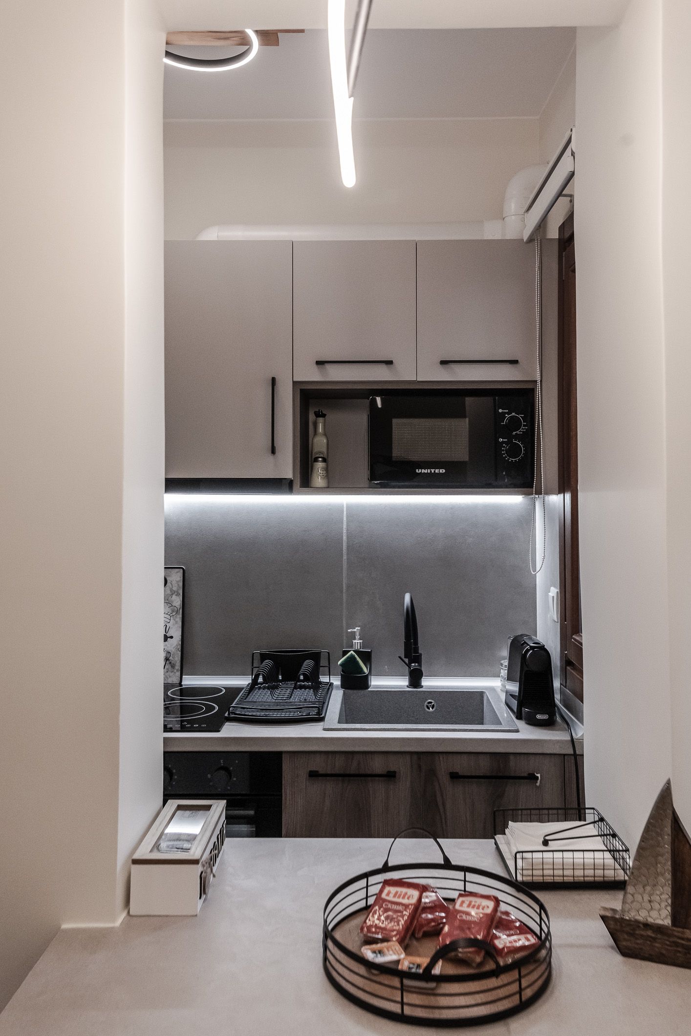 Narrow modern kitchen with white cabinets, black countertop, under-cabinet lighting, and a tray of food on a table